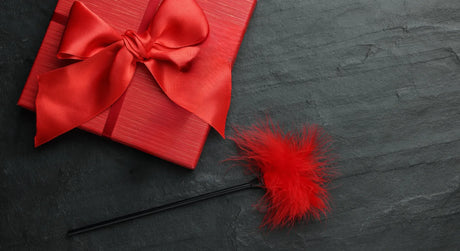 Red gift box with satin bow and feather tickler on dark background, symbolizing sensual BDSM accessories